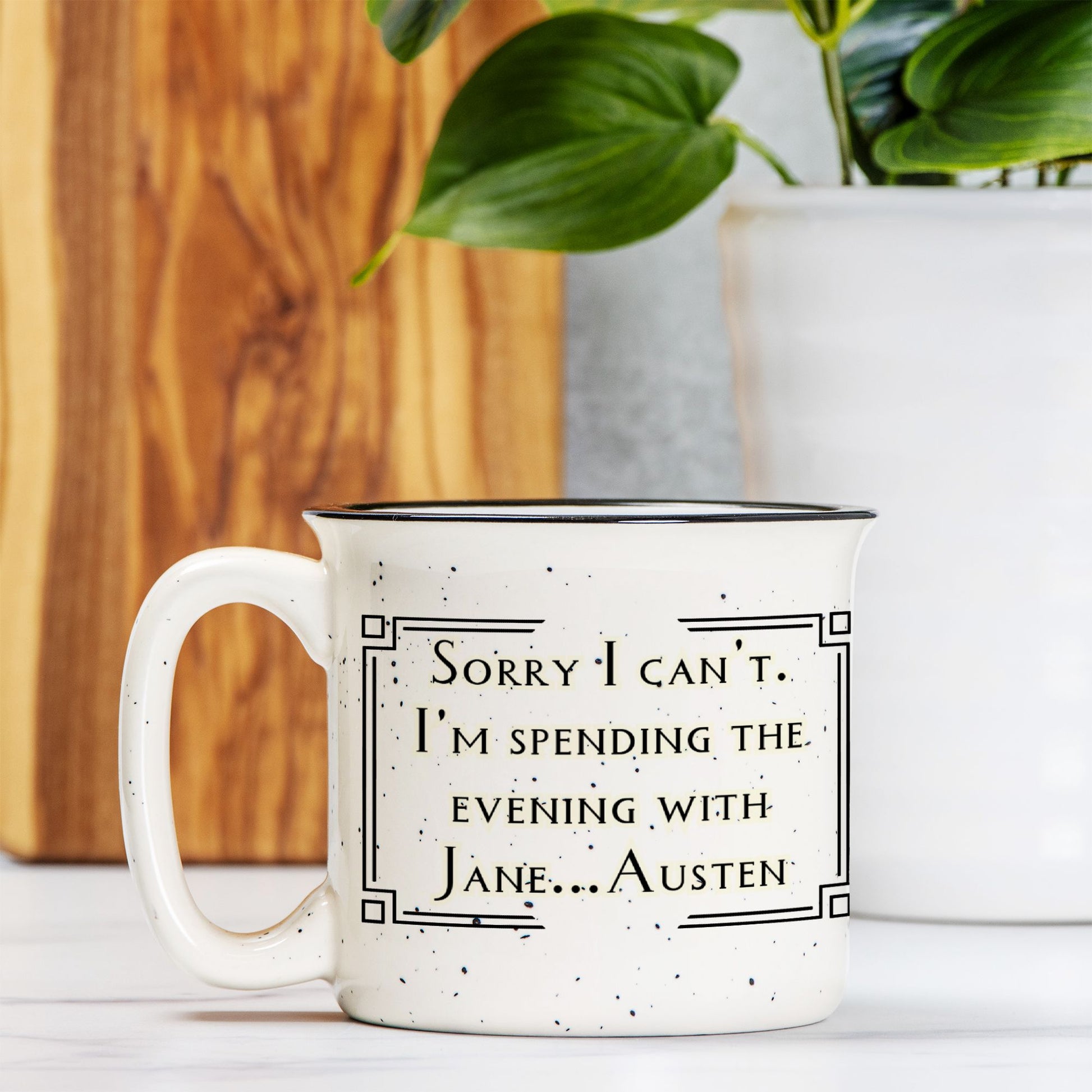 White mug with text 'Sorry I can't. I'm spending the evening with Jane Austen' on a wooden surface with a plant in the background.
