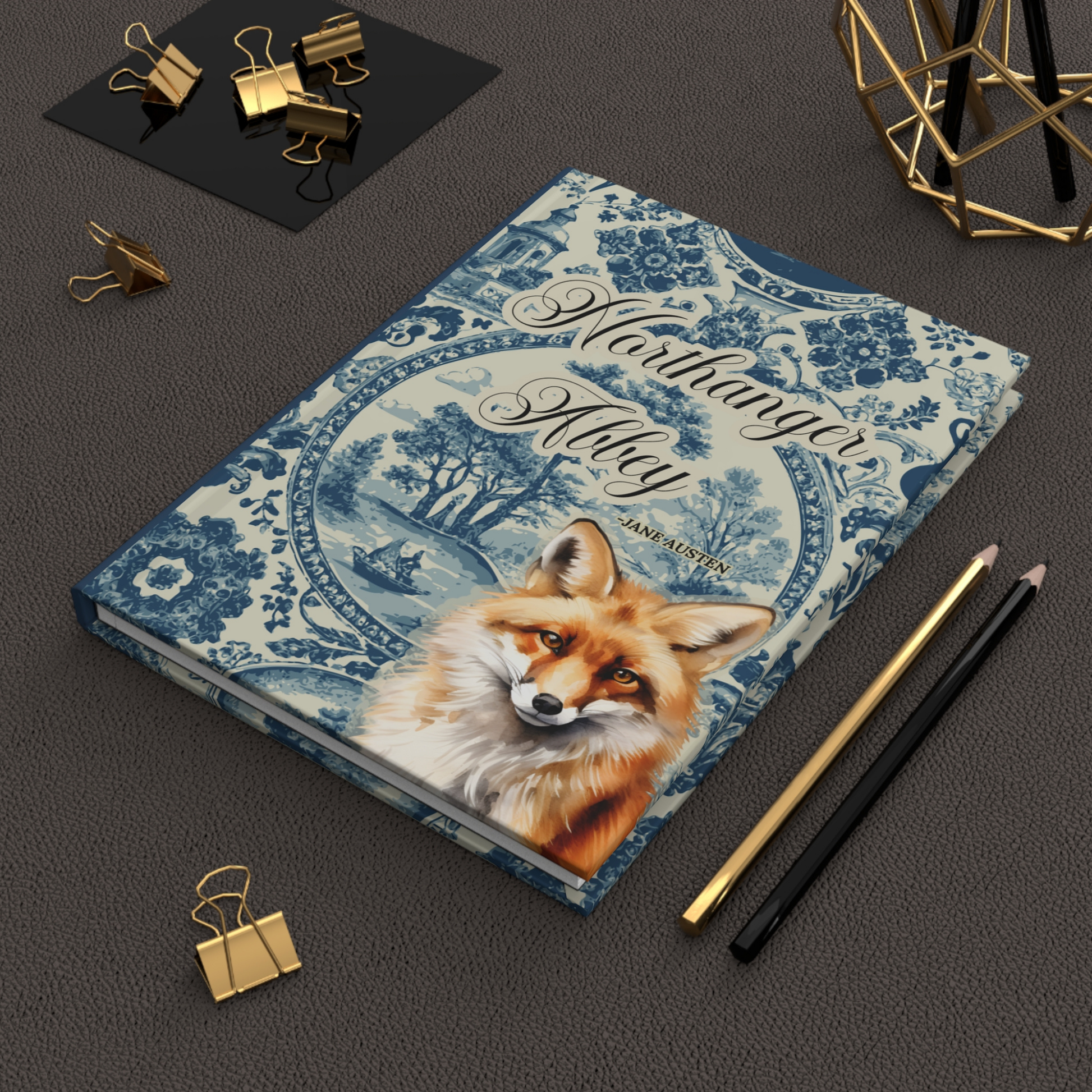 Notebook with fox illustration and 'Northanger Abbey' text on a dark surface with stationery items.