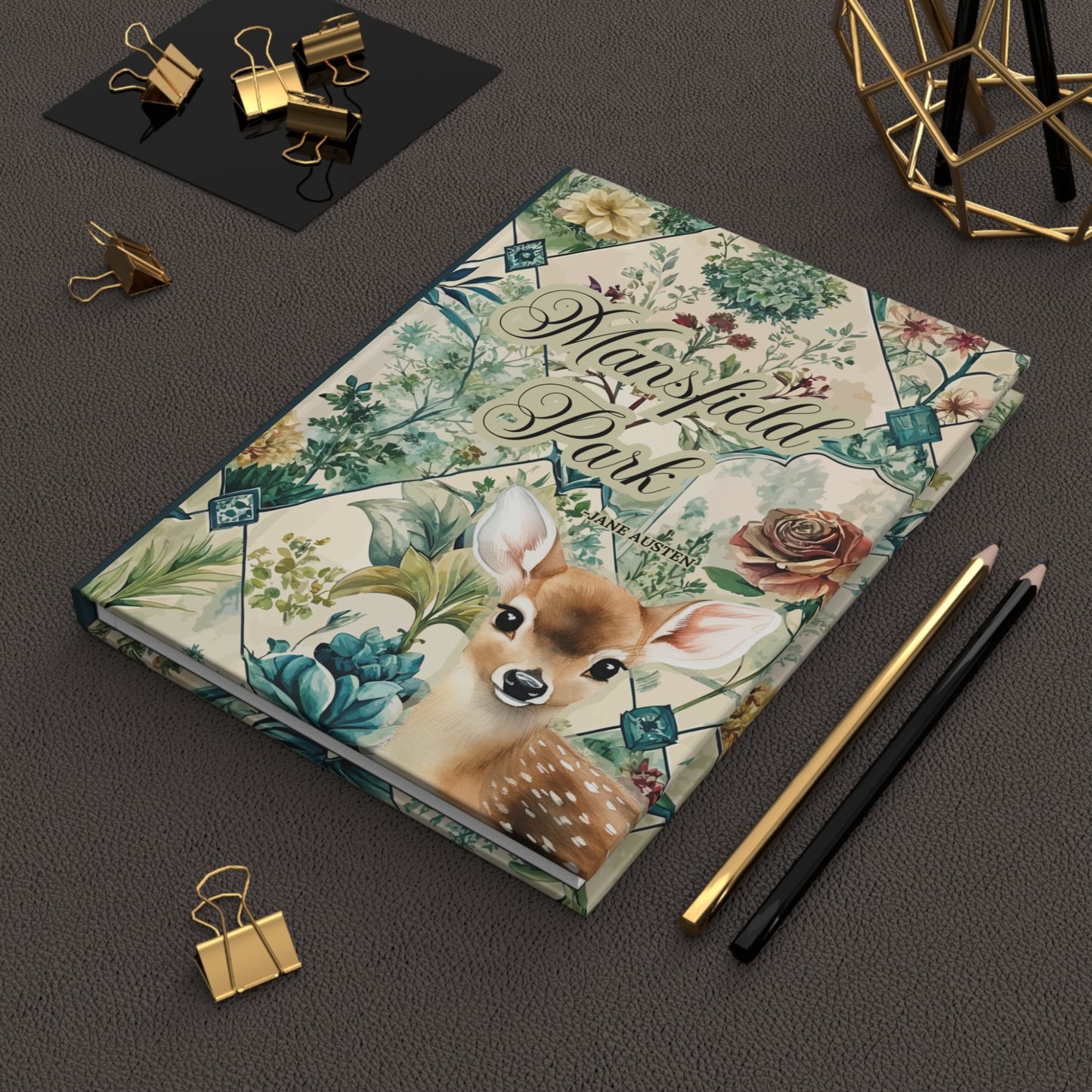 Notebook with floral and deer design on a dark surface with stationery items.