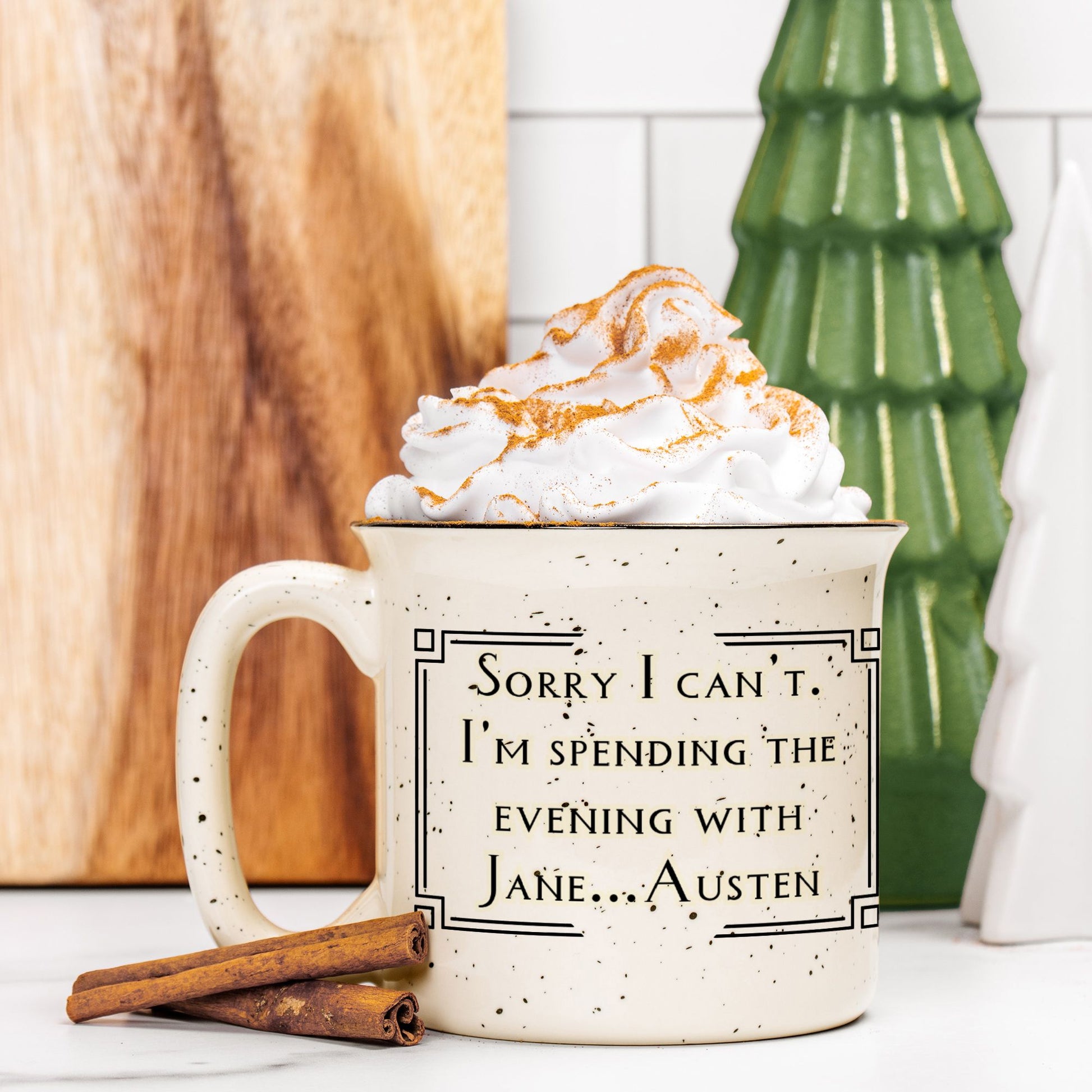 Mug with text 'Sorry I can't. I'm spending the evening with Jane Austen' filled with hot chocolate and whipped cream, surrounded by cinnamon sticks and a decorative Christmas tree.