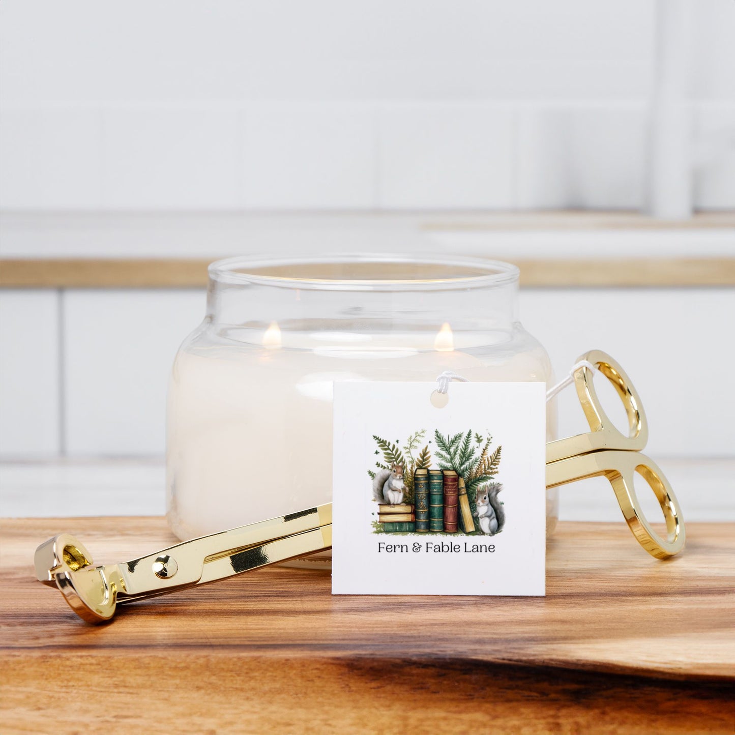 Glass candle jar with a lit candle, gold wick trimmer  and a decorative card on a wooden surface.