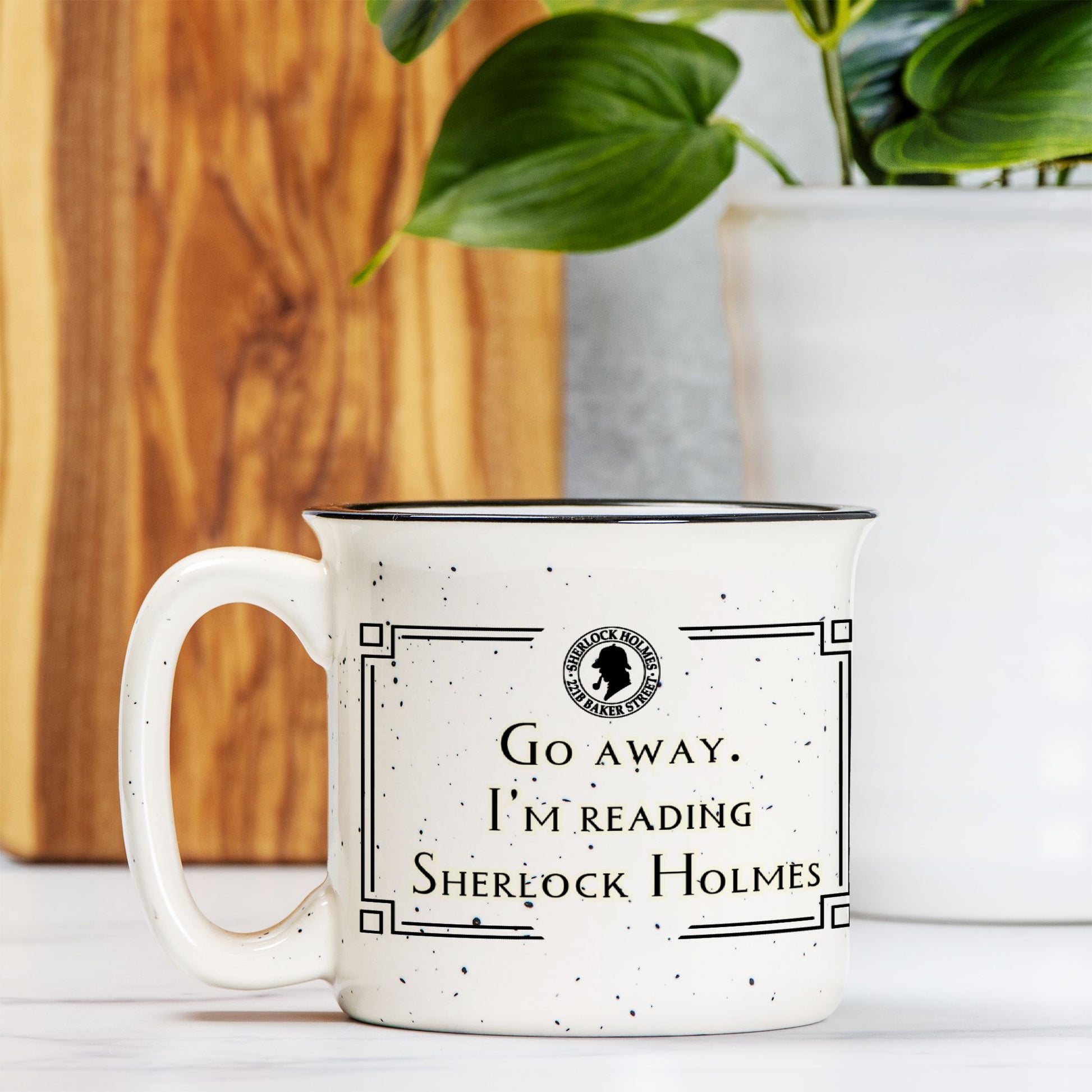White mug with text 'Go away. I'm reading Sherlock Holmes' on a wooden surface with a plant in the background.