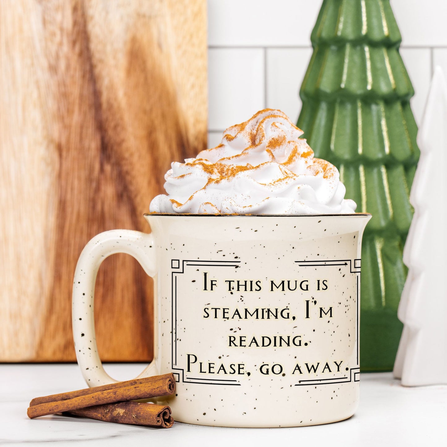 Mug with whipped cream and a humorous quote, surrounded by cinnamon sticks and a decorative tree.